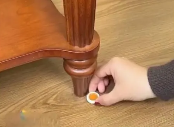 Hand applying a small white container with an orange substance to a wooden furniture leg.