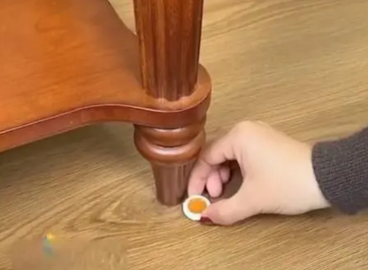 Hand applying a small white container with an orange substance to a wooden furniture leg.
