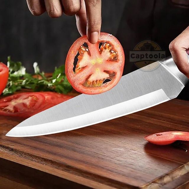 Sliced tomato held above a chef's knife on a wooden cutting board with a dark background.